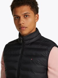 Colete puffer preto com fecho frontal e gola alta. Apresenta costura horizontal e um pequeno logótipo no peito. Usado sobre um suéter rosa.