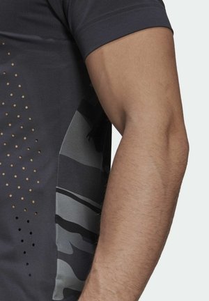 Dunkelgraues Sportshirt mit kurzen Ärmeln, das ein Camouflage-Muster in Grautönen und Schwarz, strukturierten Stoff und perforierte Details an der Seite aufweist.