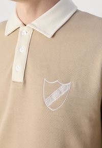 Beige pikétröja med en kontrasterande krämfärgad krage, med tre knappar och broderad logotyp på vänster bröst.