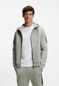 Grijze zip-up hoodie met zwarte accenten, voorzien van een trekkoord capuchon en groene rand. Gecombineerd met een witte crew neck shirt. Logo weergegeven.