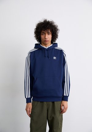 Marineblå hettegenser med hvit tre-stripes design på skuldrene, frontlompe med kenguro-design, snorhette og brodert logo på brystet.