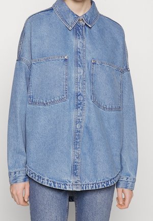 Chemise en jean bleu clair surdimensionnée avec deux grandes poches avant et des poignets boutonnés, portée sur un jean bleu assorti.