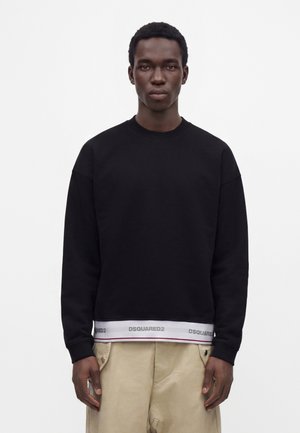 Junger Mann trägt schwarzen Sweatshirt mit weißem DSQUARED2-Logo-Band am Saum und beigen Hosen vor weißem Hintergrund.