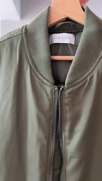 Olive green bomber jacket with a ribbed collar, featuring a sturdy zip closure and smooth, shiny fabric. Label reads "Pier One."
