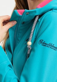 Hand justerar dragkedjan på en turkos jacka med rosa fleecefoder och svart broderad text på ärmen.