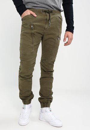 Mężczyzna ubrany w oliwkowozielone spodnie dresowe typu cargo z kieszeniami na zamek, szarą i czarną bluzę z długim rękawem oraz białe trampki, stojący na jednolitym tle.