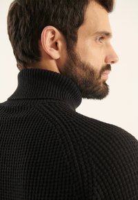 Maglione nero lavorato a maglia con un alto colletto a costine. La ricca texture e il motivo a maglia prominente sono visibili, evidenziando un design aderente.