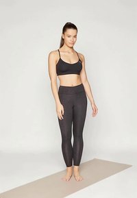 Fekete sportmelltartó és mintás fekete leggings, testhezálló kialakítással. Simaság, vékony pántokkal és magas derékkal. Egy szőnyegen áll.