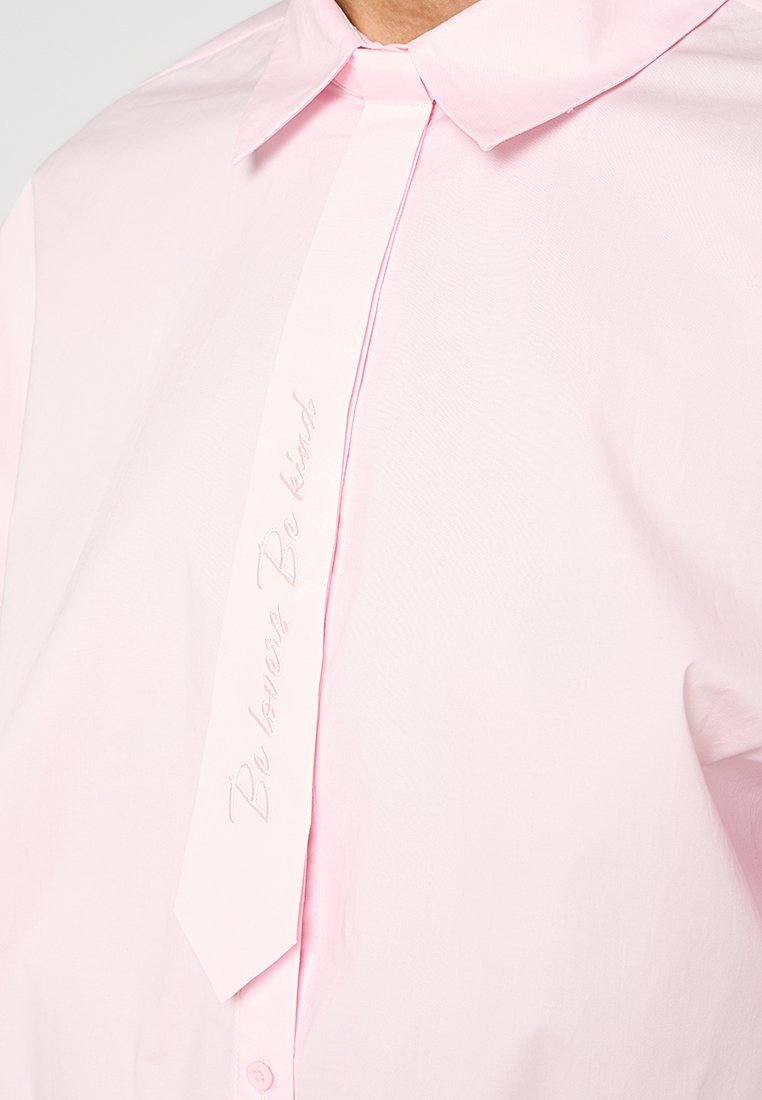Camisa de color rosa claro hecha de tela suave, con cuello puntiagudo y un detalle central de lazo con "Sé amable" bordado en cursiva.