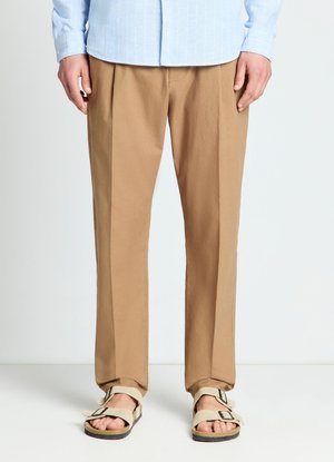 Uomo che indossa pantaloni beige a gamba dritta, camicia azzurro chiaro a maniche lunghe e sandali beige con fibbia, in piedi su pavimento bianco.