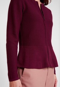 Frau trägt eine strukturierte, zum Reißverschluss gesteppte Pflaumen-Cardigan in Kombination mit hellrosa Hose, die den Stoff und die Passform der Cardigan hervorhebt.