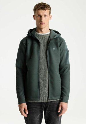 Junger Mann mit lockigem Haar, der eine dunkelgrüne Kapuzenjacke mit Reißverschluss über einem grauen Strickpullover und dunklen Jeans trägt, steht vor weißem Hintergrund.