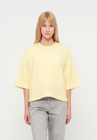 Dzeltena, pārizmēra džemperis ar īsām piedurknēm, apaļu kakla izgriezumu un mīkstu tekstūru, kombinēts ar pelēkiem džinsiem. Vienkārša, brīva silueta.