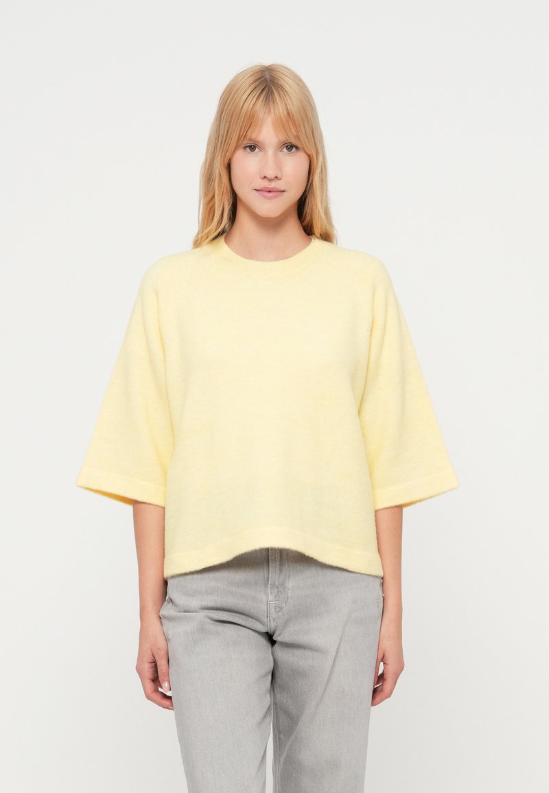 Dzeltena, pārizmēra džemperis ar īsām piedurknēm, apaļu kakla izgriezumu un mīkstu tekstūru, kombinēts ar pelēkiem džinsiem. Vienkārša, brīva silueta.