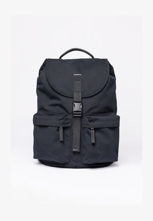 Black fabric backpack with rounded flap, adjustable strap, front pockets, and a top handle. Features smooth texture and minimal branding.