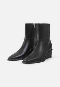 Černé kožené kotníkové boty s klínovým podpatkem, špičatou špičkou a bočním zipem. Hladká textura s minimalistickým designem, sahající až k kotníku.