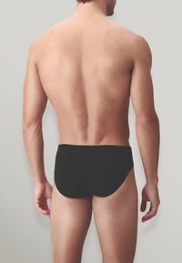 Brief neri da nuoto con una superficie liscia, vestibilità aderente e taglio basso, visti di spalle su uno sfondo neutro.