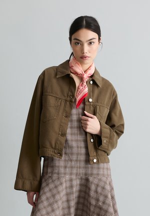 Vrouw met donker haar, gekleed in een kort bruin jasje, een geruite jurk en een roze sjaal met patroon, tegen een lichte, effen achtergrond.