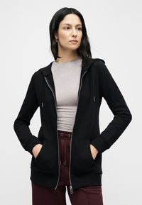 Black zip-up hoodie with a front pocket, ribbed cuffs, and a hood, worn over a grey t-shirt and paired with burgundy pants.