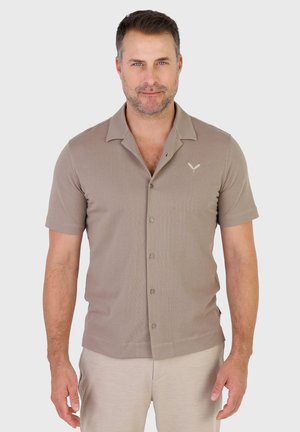 Mand med kort mørkt hår og skægstubbe iført en taupefarvet kortærmet skjorte med knapper og et lille logo på brystet, stående foran en ensfarvet baggrund.