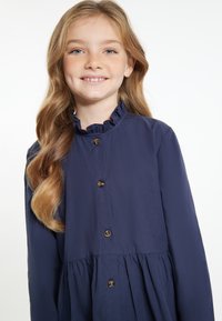 Marineblaues Hemdkleid mit Rüschenschal, langen Ärmeln und einer Taillennaht. Verfügt über runde braune Knöpfe und einen schwingenden Rock.