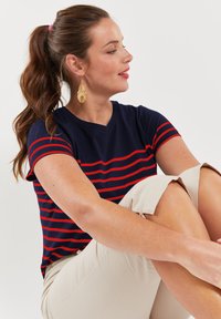 Granatowy t-shirt w paski z czerwonymi liniami, krótkie rękawy i dekolt w serek, zestawiony z beżowymi szortami. Gładki materiał i luźny krój.