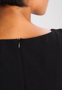 Kvinnas nacke och övre rygg synlig, klädd i en svart klänning med en stängd dragkedja vid nacklinjen, framför en enfärgad bakgrund.