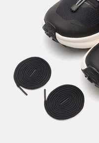 Czarne buty sportowe z teksturowanym materiałem i białą podeszwą, uzupełnione o dwa okrągłe czarne sznurowadła, wyeksponowane na białym tle.