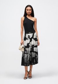 Μαύρη τοπ με έναν ώμο και εφαρμοστή μέση, συνδυασμένη με πλισέ μαύρη φούστα με μεγάλες λευκές φυλλωσιές. Το μοντέλο κρατά μια μπεζ τσάντα.