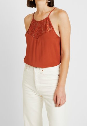 Vrouw draagt een roestrode mouwloze top met kanten rand, ingestopt in een hooggetailleerde gebroken-witte spijkerbroek met zichtbare gouden vierkante oorbellen.