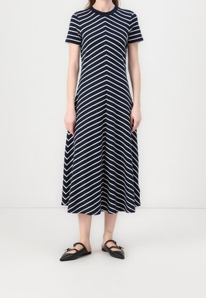 Frau trägt ein marineblaues Midikleid mit weißen diagonalen Streifen und kurzen Ärmeln, kombiniert mit schwarzen spitzen flachen Schuhen mit Riemen.