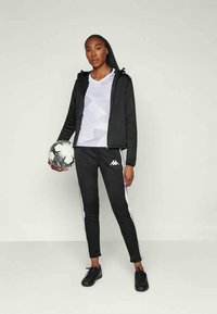 Schwarze Zip-Jacke über einem weißen T-Shirt mit geometrischem Muster, kombiniert mit schwarzen Hosen mit weißen Seitenstreifen. Hält einen schwarz-weißen Fußball.