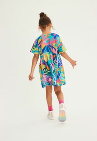 Värikäs lyhythihainen mekko, jossa on kirkas kukkakuosi sinisen, vaaleanpunaisen, keltaisen ja vihreän sävyissä, yhdistettynä valkoisiin lenkkareihin ja vaaleanpunaisiin sukkiin.