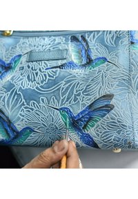 Lichtblauwe leren handtas met reliëf bloemenpatronen en met de hand geschilderde kolibries in turquoise, groen en blauw. Persoon schildert details.