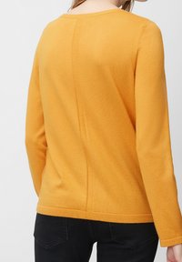 Pull en moutarde avec des manches longues, un col rond et une finition texturée. Présente une couture centrale dans le dos et un ourlet côtelé.