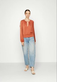 Mulher vestindo uma blusa de mangas longas laranja queimado com decote em gota, calças de ganga de cintura alta azul claro e sapatos de salto bloco abertos na frente, de pé contra um fundo branco.