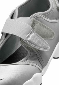 Zbliżenie na szary but wsuwany z elastycznym paskiem i regulowanym zapięciem na rzep nad wyściełanym otworem oraz białą podeszwą.