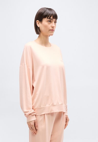 Γυναίκα με κοντά καστανά μαλλιά, φορώντας άνετη ανοιχτό ροζ μακρυμάνικη μπλούζα και παντελόνι στο ίδιο χ ρώμα, στέκεται μπροστά σε απλό ανοιχτό φόντο.