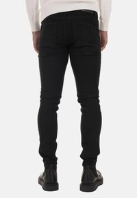 Skinny black denim jeans with a smooth texture, featuring angled back pockets and a mid-rise waistband. Worn with black ankle boots.
