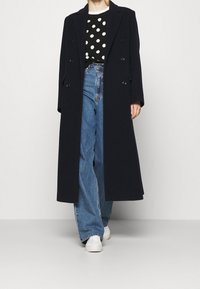 Person wearing a long black double-breasted coat, black and white polka dot top, blue jeans, and white sneakers, standing against a white wall.