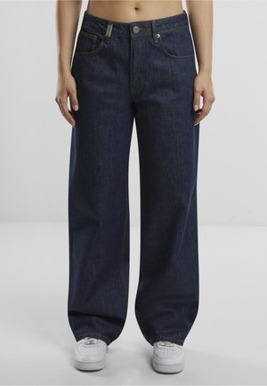 Osoba nosząca luźne ciemnoniebieskie dżinsy i białe trampki, stojąca z rękami swobodnie opuszczonymi bokiem na jednolitym tle.