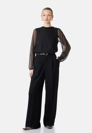 Vrouw draagt zwarte broek met wijde pijpen en een zwarte top met doorschijnende, patroon lange mouwen, taille met ceintuur en schoenen met slangenhuidprint.