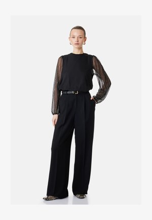 Femme portant un pantalon noir à jambes larges et un haut noir avec des manches longues transparentes et à motifs, taille ceinturée et chaussures à motif peau de serpent.