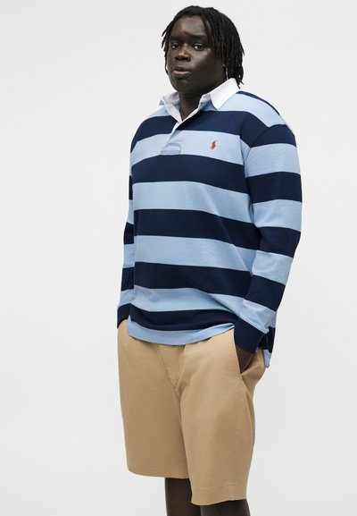 Férfi, kék és tengerészkék csíkos hosszú ujjú rögbimezben és bézs rövidnadrágban, zsebében tartott kézzel, egyszínű háttér előtt áll.