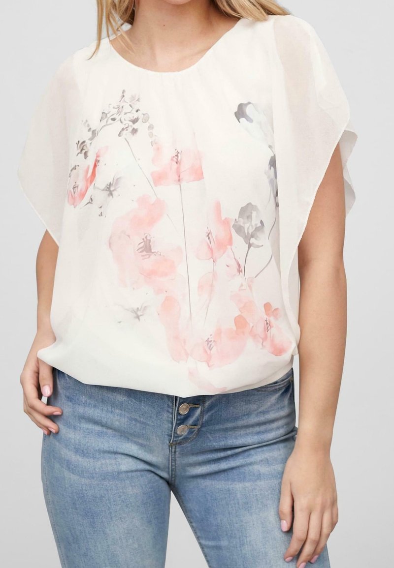 Femme portant un chemisier blanc à motif floral rose pâle et gris et un jean taille haute bleu clair avec braguette à boutons.