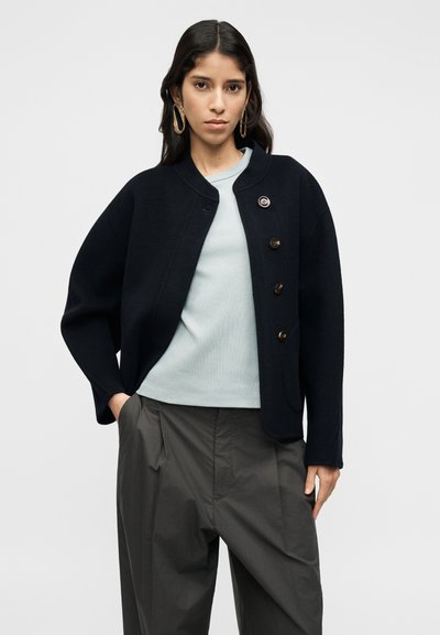 Μπλε σκούρο μάλλινο σακάκι με στρογγυλή λαιμόκοψη, τέσσερα καφέ κουμπιά, πλαϊνές τσέπες και άνετη εφαρμογή, φορεμένο πάνω από ανοιχτό μπλε μπλούζα και γκρι παντελόνι.