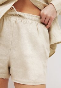 Beige linen shorts with an elastic waistband and drawstring. Smooth texture with side pockets and a simple hem finish.