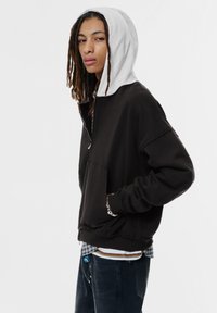 Felpa nera con zip e cappuccio grigio chiaro, dotata di tasca frontale e polsini a coste. Il modello indossa un look a strati con una camicia a quadri sotto.