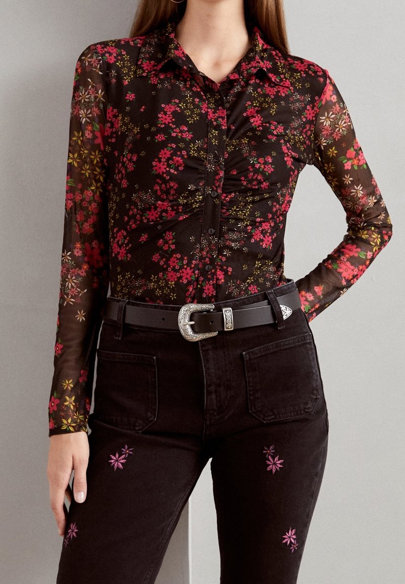 Femme portant un chemisier floral noir transparent à manches longues avec un devant froncé, associé à un jean noir brodé de fleurs roses et une ceinture noire.