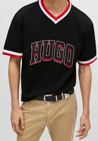 Svart kortärmad t-shirt med en röd och vit V-ringning, med djärva "HUGO"-bokstäver i röd och svart, bärs tillsammans med beiga byxor.